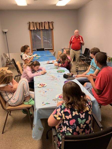 Several youths at a table with crafts and an older man standing and speaking